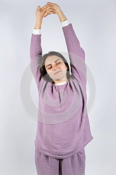 Young brunette woman in muslin lavender pajamas sleep clothes stretches by raising her arms up and closing her eyes