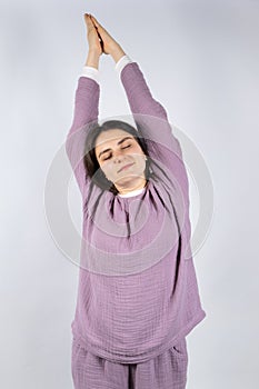 Young brunette woman in muslin lavender pajamas sleep clothes stretches by raising her arms up and closing her eyes