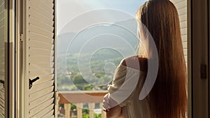 Young brunette woman opens window blinds at her villa and walking out on the terrace or balcony. Concept of summertime