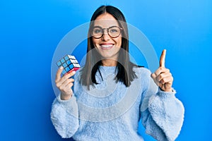 Young brunette woman playing colorful puzzle cube intelligence game smiling with an idea or question pointing finger with happy