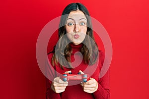 Young brunette woman playing video game holding controller puffing cheeks with funny face