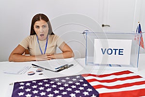 Young brunette woman at political election sitting by ballot scared and amazed with open mouth for surprise, disbelief face