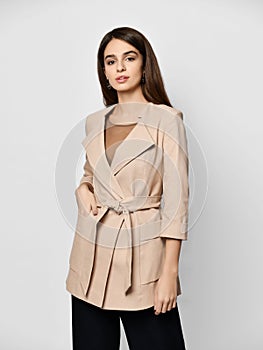 Young brunette woman is posing in stylish beige office suit jacket with large collar, big pockets and cloth belt