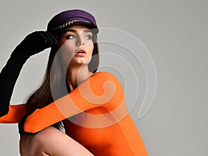 Young brunette woman in purple peaked cap beret in black gloves and orange blouse sitting and looking at the corner