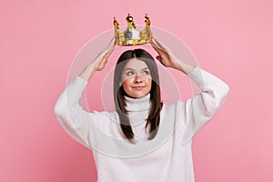 Young brunette woman putting on golden crown, looking with arrogance and smile, privileged status.