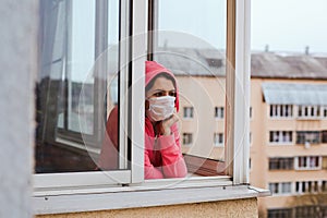 A young brunette woman is quarantined in her apartment, stands in a medical mask on the balcony and looks out the window