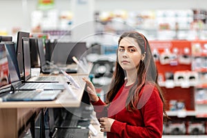 A young brunette woman in a red sweatshirt chooses a laptop in the store. Concept of buying digital goods