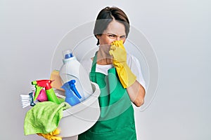 Young brunette woman with short hair wearing apron holding cleaning products smelling something stinky and disgusting, intolerable