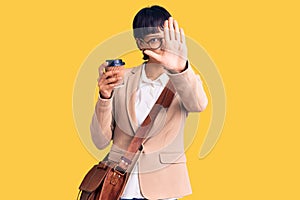 Young brunette woman with short hair wearing business bag and drinking coffee with open hand doing stop sign with serious and