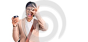 Young brunette woman with short hair wearing business bag and drinking coffee smiling happy doing ok sign with hand on eye looking