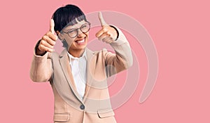 Young brunette woman with short hair wearing business jacket and glasses approving doing positive gesture with hand, thumbs up