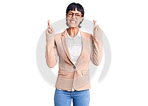 Young brunette woman with short hair wearing business jacket and glasses gesturing finger crossed smiling with hope and eyes