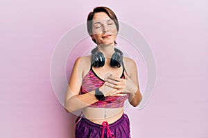 Young brunette woman with short hair wearing sportswear and headphones smiling with hands on chest with closed eyes and grateful