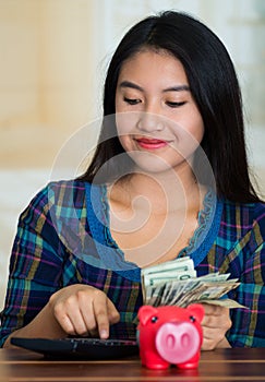 Young brunette woman sitting down facing camera, using calculator and holding stack of dollar bills, pink piggy bank on