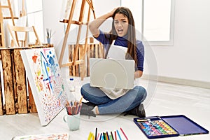 Young brunette woman sitting on the floor at art studio with laptop stressed and frustrated with hand on head, surprised and angry