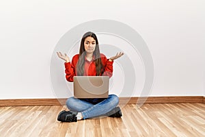 Young brunette woman sitting on the floor at empty room with laptop clueless and confused expression with arms and hands raised
