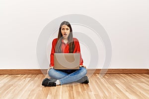 Young brunette woman sitting on the floor at empty room with laptop scared and amazed with open mouth for surprise, disbelief face