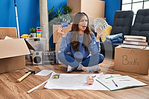 Young brunette woman sitting on the floor at new home holding savings clueless and confused expression