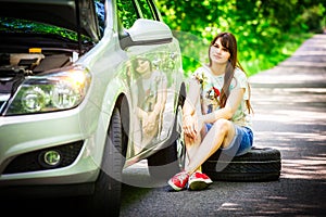 Young brunette woman sitting near a silver car on the roadside with a broken wheel