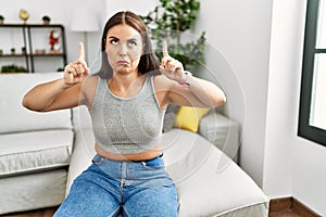 Young brunette woman sitting on the sofa at home pointing up looking sad and upset, indicating direction with fingers, unhappy and