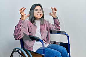 Young brunette woman sitting on wheelchair shouting frustrated with rage, hands trying to strangle, yelling mad