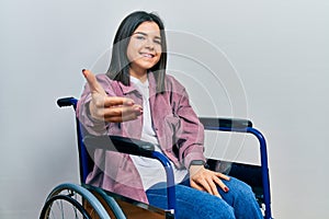 Young brunette woman sitting on wheelchair smiling friendly offering handshake as greeting and welcoming