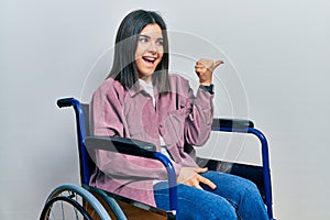 Young brunette woman sitting on wheelchair smiling with happy face looking and pointing to the side with thumb up