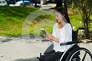 Young brunette woman sitting in wheelchair smiling with positive attitude, using mobile phone, outdoors environment