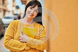 Young brunette woman smiling happy holding book leaning on the wall