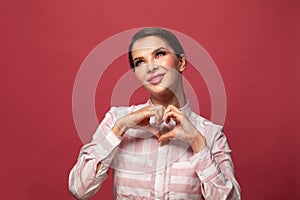 Young brunette woman smiling and showing a heart shape with hands on red background