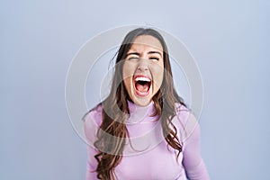 Young brunette woman standing over blue background angry and mad screaming frustrated and furious, shouting with anger