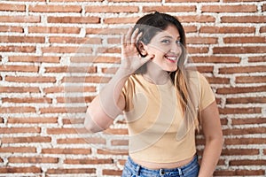 Young brunette woman standing over bricks wall smiling with hand over ear listening an hearing to rumor or gossip