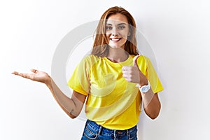 Young brunette woman standing over isolated background showing palm hand and doing ok gesture with thumbs up, smiling happy and