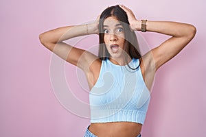 Young brunette woman standing over pink background crazy and scared with hands on head, afraid and surprised of shock with open