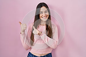 Young brunette woman standing over pink background pointing to the back behind with hand and thumbs up, smiling confident
