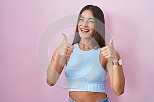 Young brunette woman standing over pink background success sign doing positive gesture with hand, thumbs up smiling and happy