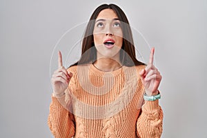 Young brunette woman standing over white background amazed and surprised looking up and pointing with fingers and raised arms