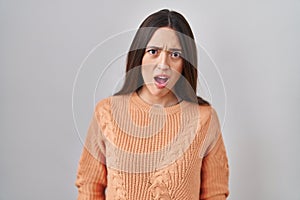Young brunette woman standing over white background in shock face, looking skeptical and sarcastic, surprised with open mouth