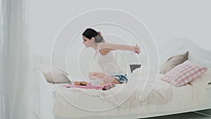 Young brunette woman stretches on a bed. Beautiful girl using laptop during breakfast. Drinking juice and dancing.