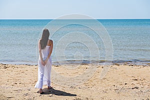 Young brunette woman in summer white dress standing on beach and looking to the sea.