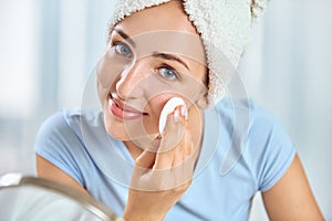 A young brunette woman with a towel wrapped round her head cleansing her skin with a cotton pad