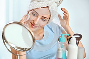 A young brunette woman with a towel wrapped round her head cleansing her skin with a cotton pad