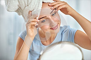 A young brunette woman with a towel wrapped round her head examining her face in the round mirror