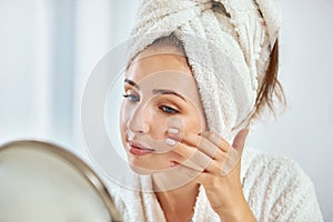 A young brunette woman with a towel wrapped round her head examining her face in the round mirror