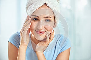 A young brunette woman with a towel wrapped round her head touching her cheeks by her both hands