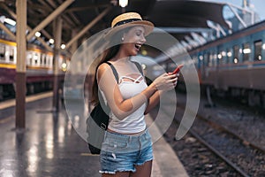 Young brunette woman traveler laughs while playing a mobile phone in train station platform.