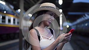 Young brunette woman traveler laughs while playing a mobile phone in train station platform