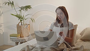 Young brunette woman uses laptop while lying in bed. A girl in a white T-shirt is typing on a laptop keyboard on her lap