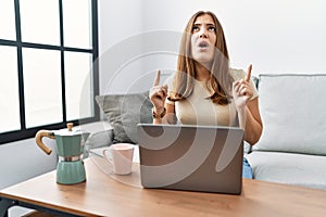 Young brunette woman using laptop at home drinking a cup of coffee amazed and surprised looking up and pointing with fingers and