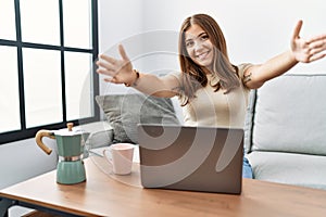 Young brunette woman using laptop at home drinking a cup of coffee looking at the camera smiling with open arms for hug